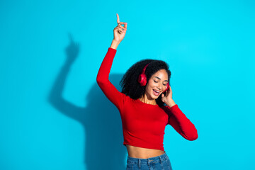 Young stylish woman enjoying music with headphones on a vibrant blue background, wearing a red top, dancing happily