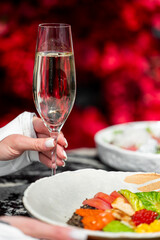 A hand elegantly holding a champagne flute with a vibrant red background and a colorful plate of food. A stylish and celebratory moment captured in detail