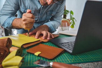 Leather craftsman working with leather using a stitching groover and laptop in his workshop