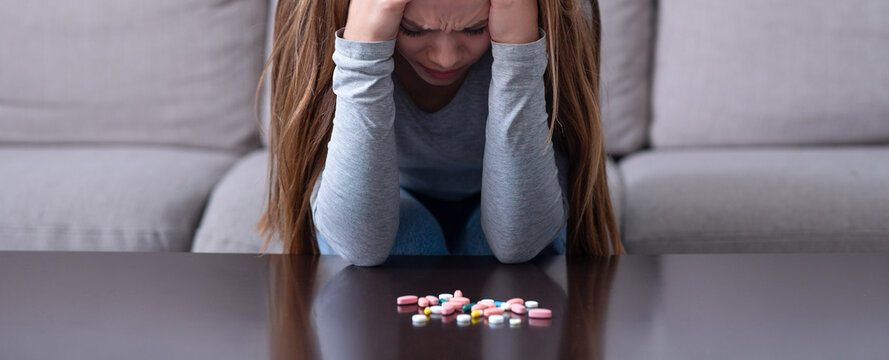 Depressed young woman sitting head in hands near pile of tablets, planning to commit suicide at home. Desperate millennial lady with emotional problems overdosing on meds, killing herself