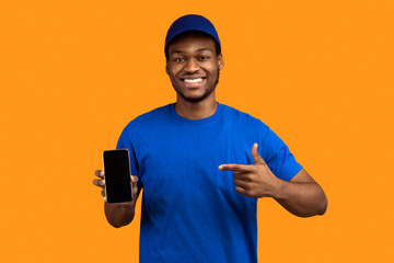 Recommending Service. Smiling african american delivery guy holding smartphone with blank screen for mockup, wearing blue cap and uniform, pointing at black empty monitor isolated on orange background