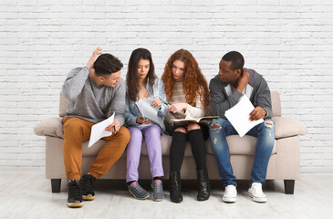 Fototapeta premium Multiethnic young students studying, preparing for exam, sitting on sofa in living room against white brick wall, studio shot