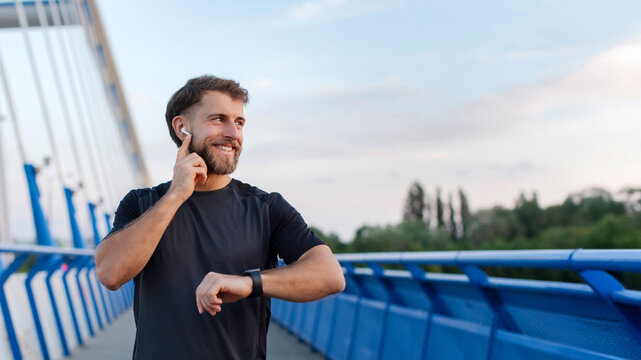 Handsome athlete man training outdoors on bridge, runner using earphones and smartwatch for health data and statistics, panorama, free space - Powered by Adobe
