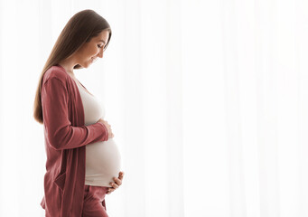 Profile Portrait Of Smiling Pregnant Woman Tenderly Embracing Belly Against Window At Home, Happy Expectant Mother Enjoying Her Healthy Pregnancy And Awaiting Time, Side View With Copy Space © Prostock-studio