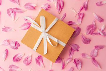 Closeup of gift box with white ribbon on scattered peony petals and pink background..