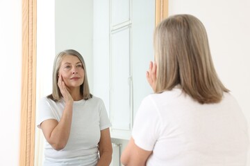 Beautiful senior woman looking at herself in mirror indoors