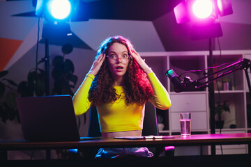 Young woman streaming at night in a neon-lit room with microphone and computer showing excitement...