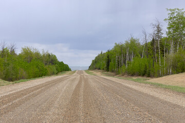 Single lane dirt country road with gravel in the rural area, green trees on sides, spring blue sky and rain in the horizon.