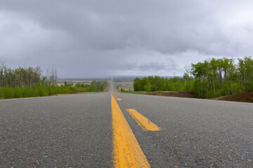 Highway with black asphalt and yellow lines in the rural area, green trees on sides, cloudy spring blue sky.