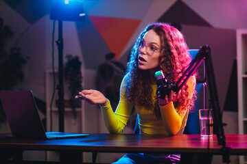 Young woman blogger streaming online using microphone and laptop in neon lit studio