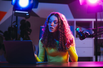 Young woman livestreaming with bright neon lights using a laptop and microphone in a modern indoor...