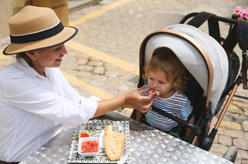 Mother Feeding Child at Outdoor Caf? Table with Stroller Nearby on Sunny Day