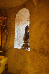 A stone statue of a robed figure stands behind a barred arched window, illuminated by natural light...