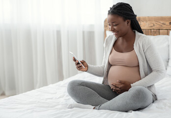 Smiling Black Pregnant Woman Sitting With Smartphone On Bed At Home, Looking At Cellphone Screen...