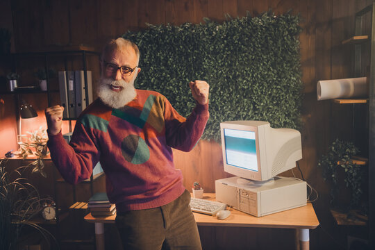 Joyful senior man celebrating in a cozy working space with classic computer setup embodying technology and retro style energy