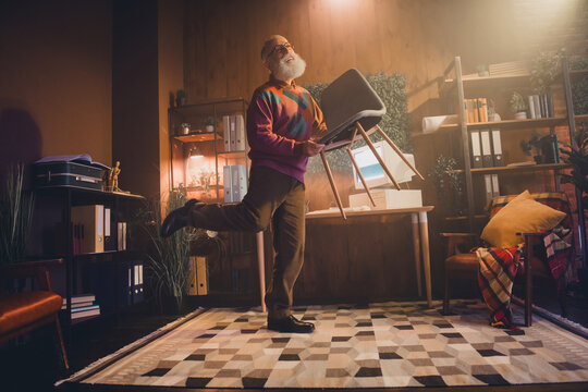 Elderly man enthusiastically holding a chair in a cozy home office setting filled with technology and bookshelves