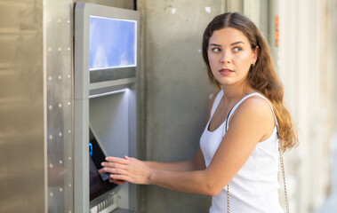 Young girl looking alert and slightly tense while standing at outdoor city ATM, shielding keypad...