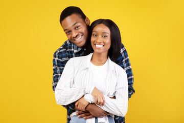 Fototapeta premium Happy romantic date or self-isolation together. Cheerful young african american male and female in casual, hugging and looking at camera, isolated on yellow background, studio shot, copy space