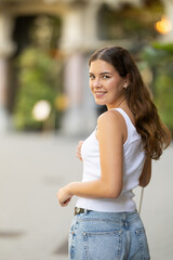 Young woman in early 20s wearing white tank top and blue jeans gently smiling and turning back...