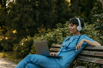 Doctor wearing headphones and using laptop while relaxing on bench in park