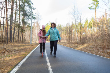 Little girl riding scooter while walking with her grandmother in the forest on a sunny day
