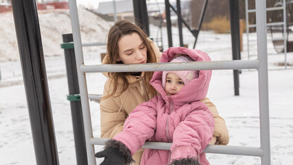 Obraz premium Young mother and a little girl in warm outerwear walk in the park in the children's playground in winter.