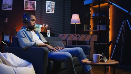 Black teleworker putting headset to listen to webinar discussion, participates at an online lesson on laptop to improve his skills. Young person enjoying meeting from his living room. Camera A.