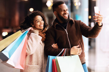 It's Shopping Time. Happy black man and woman taking selfie standing with colorful bags near...