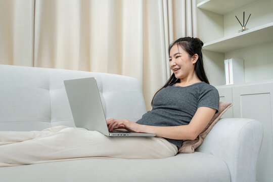 Asian freelance woman using laptop to receive email of order from client and lying on the couch to checking product on stock