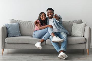 Happy laughing relaxed millennial African American woman and man watching tv in living room interior, holding remote control and hugging. Smiling couple enjoying film and movie evening together