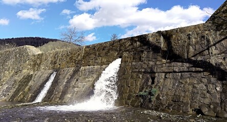 A river dam in a wilderness area. Water flow. Warm season. Bright picture. Cloudy sky. A splash. Foam. Cover. Wallpaper. Picture. Background