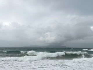 Overcast sky above the ocean with waves crashing on the shore and distant land visible on the horizon