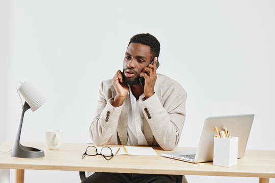 Professional man talking on mobile phone while working at minimalist office desk with laptop, glasses, and stationery; workspace with clean, bright background; business communication concept.