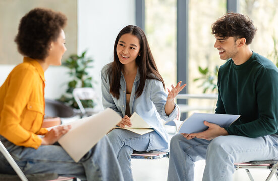 Three engaged diverse students sit in well-lit modern classroom, with one gesturing as they enthusiastically discuss the contents of their notebooks