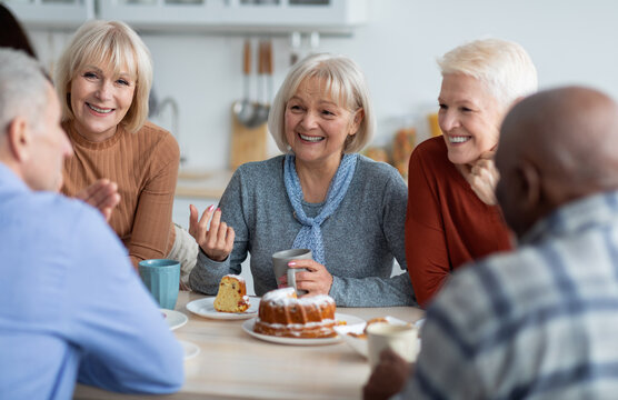Healthy and active positive senior people of different nationalities sitting at kitchen, drinking tea and eating cake together, having conversation and laughing, chilling together at nursing home