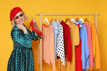 Smiling fashion stylist and rack with clothes on yellow background