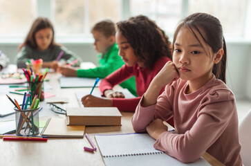 Unhappy Asian Schoolgirl Thinking Having Problem With Education Sitting While Her Diverse Classmates Learning Lesson In Modern Classroom At School. Educational Issues Concept. Selective Focus