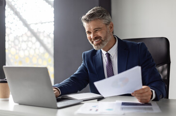 Plakat Successful middle aged entrepreneur working with laptop and papers at workplace in modern office, smiling mature businessman in suit sitting at desk and managing financial reports, copy space