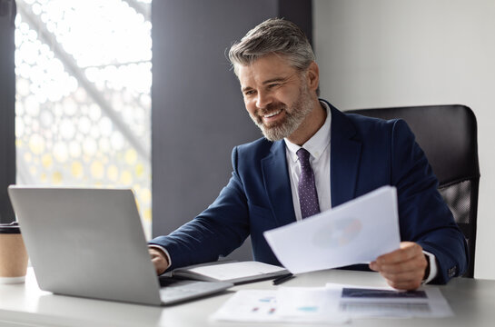 Fototapeta Successful middle aged entrepreneur working with laptop and papers at workplace in modern office, smiling mature businessman in suit sitting at desk and managing financial reports, copy space