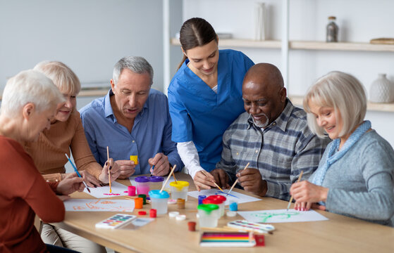 Cheerful creative multiracial senior men and women sitting around table and drawing at nursing home, doing arts and crafts together, young lady nurse assisting group of elderly people, copy space