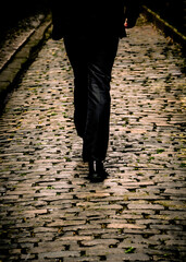 A person in a dark suit walks down a cobblestone street.