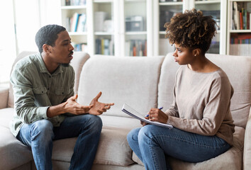 Young african american woman therapist taking notes on her upset patient at therapy session,...