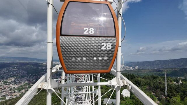 Ferris wheel cabin moving upwards, providing breathtaking views of Tbilisi cityscape, the TV Tower, and the surrounding landscape under a cloudy sky in Georgia