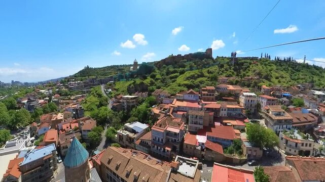 Aerial view capturing the Kura River winding through Tbilisi, with the cityscape stretching towards the horizon and the historic Narikala fortress undergoing restoration on a green hill
