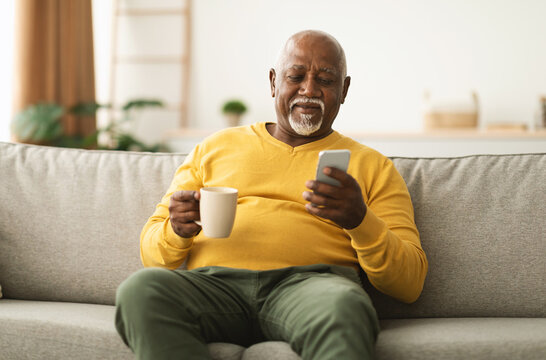 Mature African American Male Using Cellphone Browsing Internet And Texting Communicating Online In Social Media App Sitting On Sofa At Home. Senior Man Drinking Coffee And Scrolling News On Phone