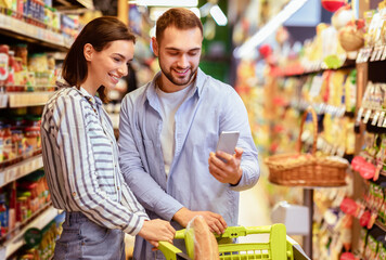 Smiling young couple shopping healthy food in supermarket, buying fresh natural products and using smartphone in store. Family at grocery using mobile phone. Husband and wife comparing prices