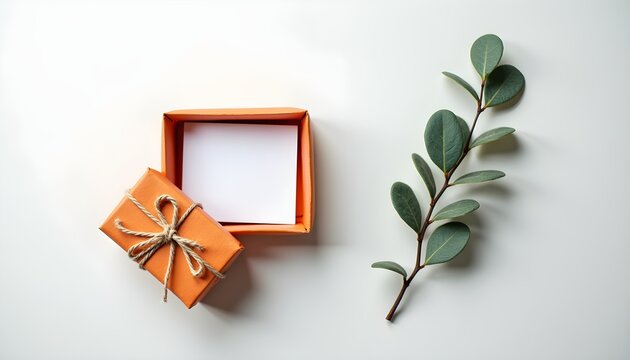 A small orange gift box with a string tied around it and a small branch of eucalyptus leaves on a light-colored surface