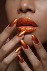 A close-up beauty shot focusing on metallic copper-orange lips and matching nail polish. The lips are painted in a glossy, metallic finish with a warm, rust-colored tone. 