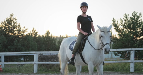 Horsewoman wearing a helmet gracefully riding a stunning white horse in a paddock at sunset,...