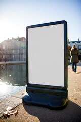 empty black-framed advertisement board standing outdoors on a sunny pavement near water with buildings and a person walking away in the background